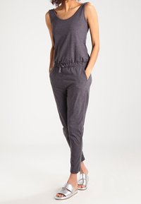 Mono sin mangas gris con cintura elástica y bolsillos laterales, combinado con sandalias deslizantes plateadas sobre un fondo claro.