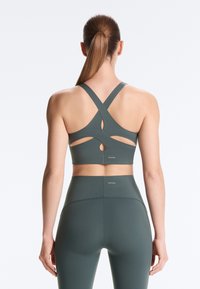 Femme avec une queue de cheval brune portant un soutien-gorge de sport vert foncé à bretelles croisées et un legging taille haute, debout de dos face à la caméra sur fond blanc.