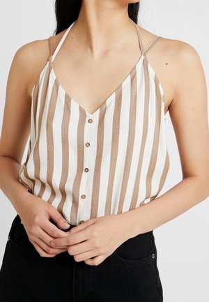 Pruhovaný bezrámový top v bílé a béžové barvě, s výstřihem do V, knoflíkovým zapínáním v přední části a tenkými ramínky. Nosený s černými kalhotami.