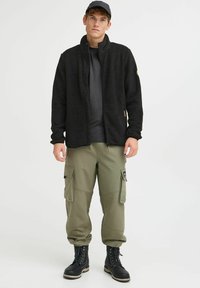 Must must vest, mille kaelusega, oliivirohelised cargo-püksid mitme taskuga ja mustad nööbiga saapaid. Lihtne disain, pehme tekstuur.