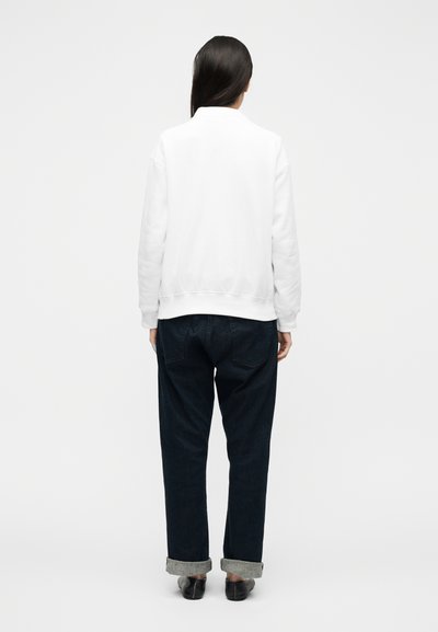 Γυναίκα όρθια πλάτη, φοράει λευκό φούτερ, σκουρόχρωμα τζιν με γυρισμένα μανίκια και μαύρα παπούτσια φόρμας, μπροστά σε απλό φόντο.