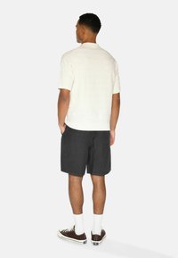 Helles beiges Kurzarmhemd mit strukturiertem Muster, kombiniert mit dunklen Anthrazit-Shorts. Braune Sneakers mit weißen Sohlen und Socken runden den Look ab.