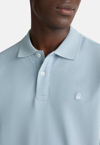 Světle modrá polo tričko vyrobené z texturované tkaniny s klasickým límečkem, dvouknoflíkovou légou a vyšitým logem chobotnice v bílé barvě.
