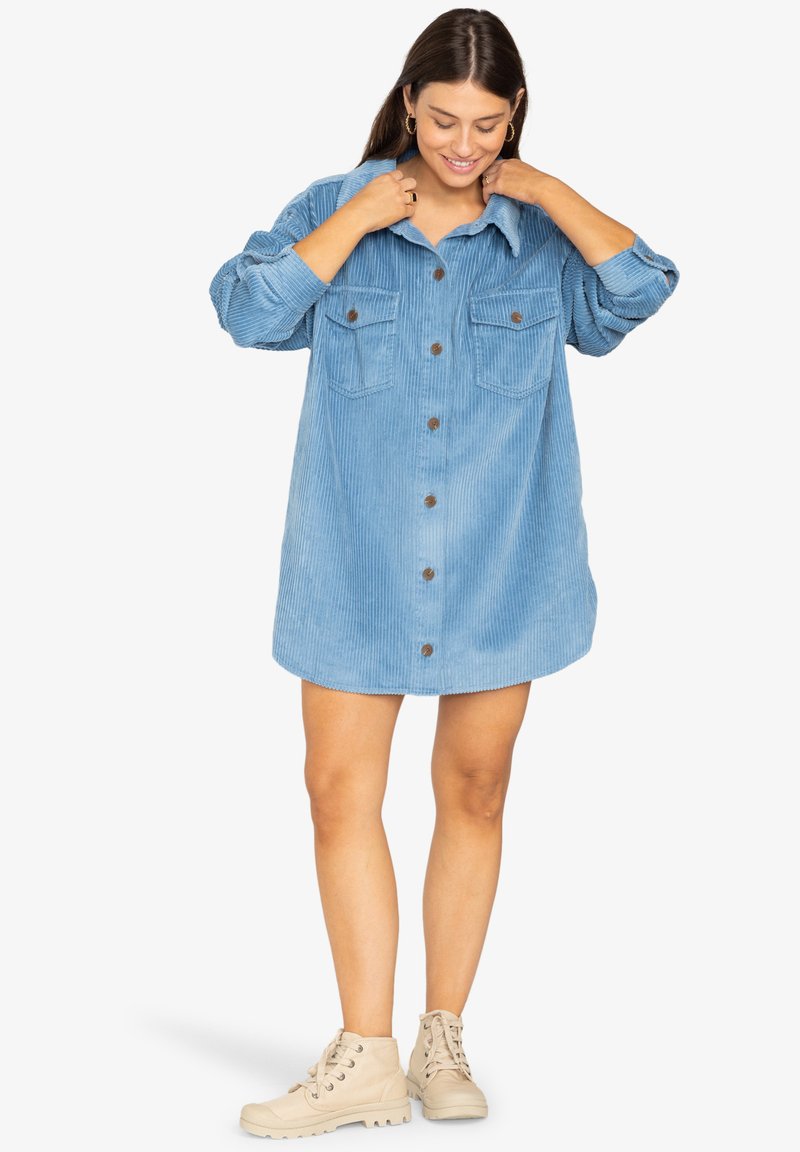Vestido camisero de pana azul con botones en la parte delantera, dos bolsillos en el pecho y mangas enrolladas. Lleva unas botas de tobillo beige.