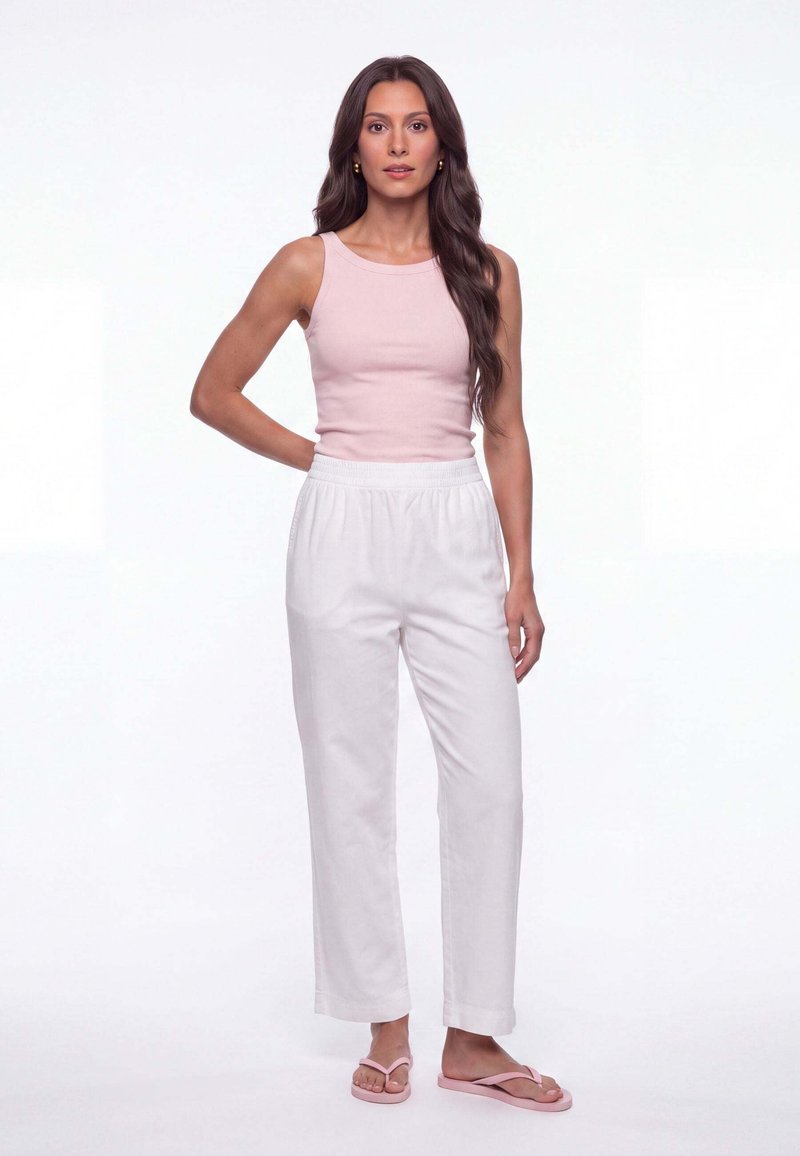Femme aux longs cheveux foncés portant un haut rose sans manches, un pantalon blanc et des tongs roses, debout avec une main sur la hanche.