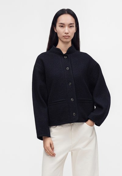 Femme portant une veste en laine noire boutonnée et un pantalon blanc, debout avec une main dans la poche sur un fond clair uni.