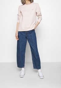 Persona che indossa un felpa a maniche lunghe rosa chiaro, jeans a gamba larga blu, sneaker alte bianche, in piedi con una mano in tasca su sfondo bianco.