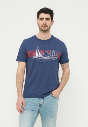 Άνδρας με σκούρα μαλλιά που φοράει μπλε μπλουζάκι με σχέδιο ιστιοφόρου και κόκκινες ρίγες, καθώς και ανοιχτόχρωμα μπλε τζιν, στέκεται με το ένα χέρι στην τσέπη.