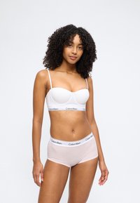 Γυναίκα που στέκεται φορώντας λευκό σουτιέν Calvin Klein και ανοιχτό ροζ σλιπ Calvin Klein μπροστά σε απλό λευκό φόντο.