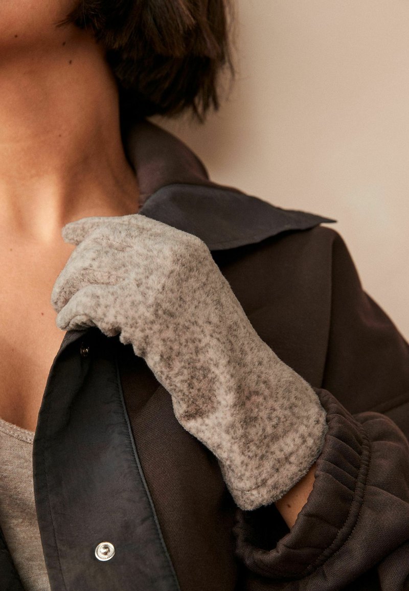 Grijze fleece handschoen met een gestructureerde afwerking, licht getailleerde vorm, gedragen op een hand. De persoon draagt een donkere jas met een kraag.