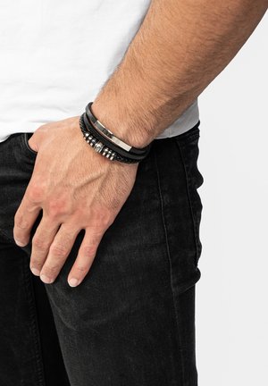 Black woven leather bracelets with metallic accents, layered on a wrist. Hand rests against dark jeans, showcasing texture and design details.