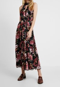 Robe maxi florale noire avec des fleurs roses et rouges, design à col halter, taille cintrée et jupe fluide. Portée avec des mocassins marron.