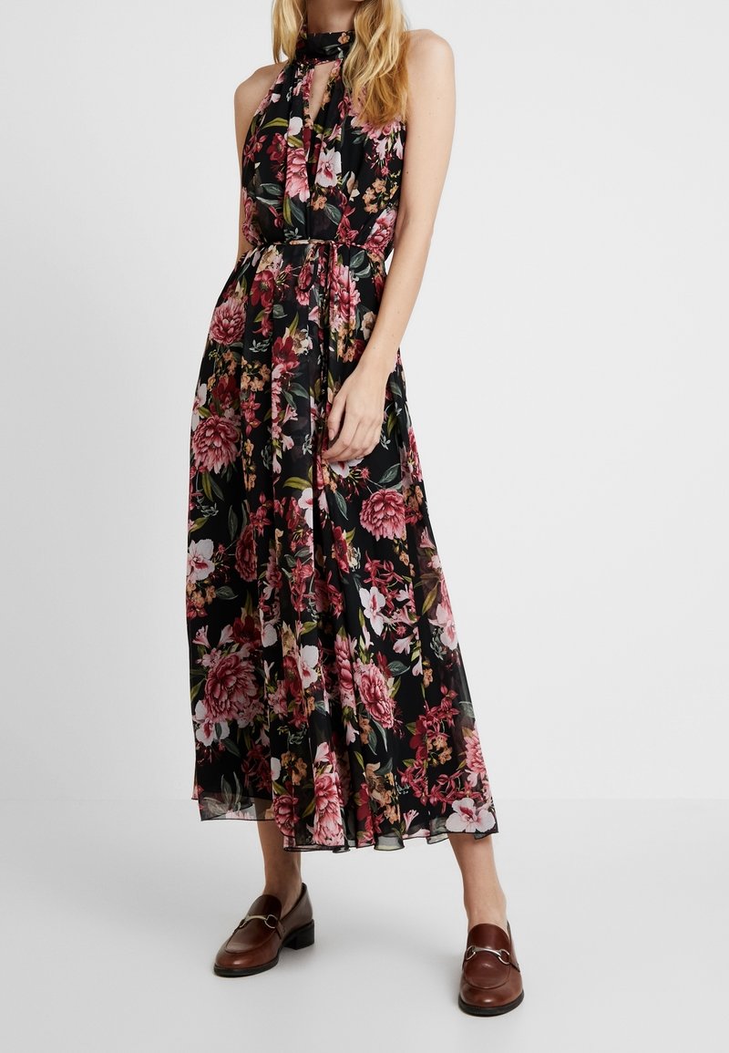 Robe maxi florale noire avec des fleurs roses et rouges, design à col halter, taille cintrée et jupe fluide. Portée avec des mocassins marron.