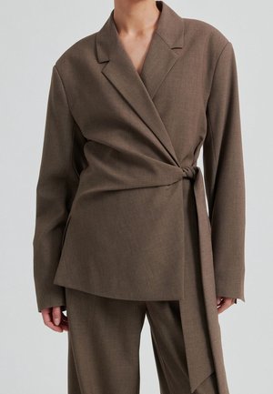 Femme portant un blazer marron noué sur le côté avec un pantalon assorti, sur un fond clair uni.