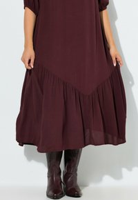 Dieprode, vloeiende jurk met een gerimpelde zoom en een V-vormig ontwerp. Gecombineerd met donkerbruine, knie-lange laarzen met gestructureerde accenten.