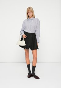 Blonde vrouw in een gestreepte blouse, zwarte flared shorts, donkere sokken tot halverwege de kuit, burgundy ballerina's, met een kleine witte handtas.