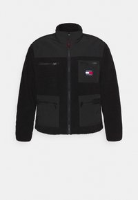 Czarna polarowa kurtka z gładką czarną łatą na górze, kieszeniami na zamek oraz naszywką z logo na klatce piersiowej. Posiada wysoki kołnierz i zapięcie na zamek z przodu.