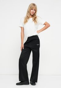 Λευκό κοντομάνικο μπλουζάκι σε συνδυασμό με μαύρο παντελόνι φόρμας με σχέδιο πλευρικής ρίγας και λογότυπο. Φορώντας μαύρα αθλητικά παπούτσια.