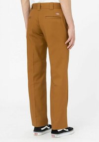 Braune Chino-Hosen mit geradem Bein. Zwei Gesäßtaschen und ein kleines Markenlabel. Kombination mit schwarz-weißen Sneakers.