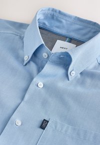 Chemise bleu clair à boutons blancs, col pointu, poche avant et étiquette de marque à l'intérieur du col et sur la poche.