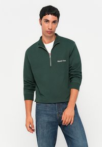Zelený pulovr na polo zip v měkké tkanině s žebrovanými manžetami a lemem, se bílým vyšívaným textem na hrudi, spárovaný s džíny.