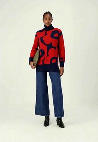 Žena v červeném a námořnickém květinovém roláku, v širokých modrých džínách, s černými kotníkovými botami, drží olivově zelenou kabelku.