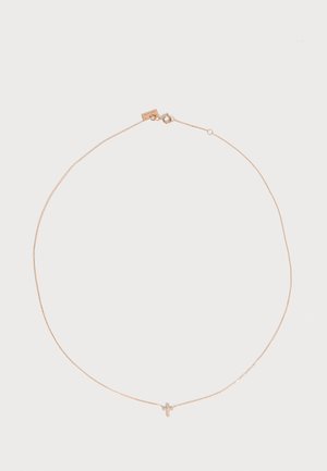 Collier fin en chaîne dorée avec un petit pendentif en forme de croix orné de pierres claires sur un fond clair uni.