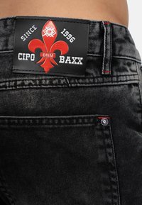 Black denim jeans with a leather patch featuring a red fleur-de-lis logo and white text. Includes red stitching and metal button details.