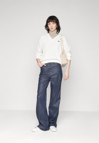 Witte V-hals trui, beige coltrui eronder, donkerblauwe wijde jeans, witte sneakers en een gestructureerde beige tas.