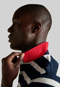 Krekls ar apkakli, ar tumši zilu un baltu svītru rakstu un sarkanu iekšējo apkakli, izgatavots no gludas auduma. Apkakli ar roku pielāgo no kreisās puses.