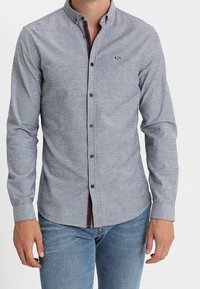 Jasnoszara koszula na guziki z długim rękawem i ostrym kołnierzykiem, z delikatną fakturą i małym czarnym logo na piersi.