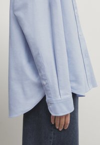 Poignet de chemise bleu clair à manches longues avec boutons porté sur un jean en denim foncé, main détendue sur le côté sur fond uni.