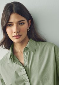 Chemise vert clair à boutons, dotée d'un col et de coutures visibles. Le modèle porte des boucles d'oreilles argentées en créole.