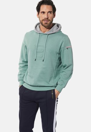 Minzgrüner Kapuzenpullover mit grauem Innenfutter, vorne mit einem strukturierten Logo und einer seitlichen Reißverschlusstasche, kombiniert mit marineblauen Trainingshosen.