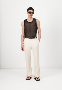 Schwarzes Mesh-Top, cremefarbene weit geschnittene Hose, schwarzer Gürtel und schwarze Flip-Flops. Das Model trägt außerdem schwarze rechteckige Sonnenbrillen.