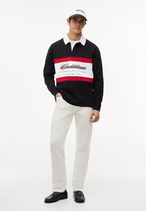 Mladý muž oblečený v čiernej, červenej a bielej dlhoruká polo košeľa s nápisom "Cadillac Formula 1 Team", bielych nohaviciach, čiernych topánkach a bielej šiltovke.