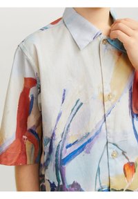 Lapsi, joka pukeutuu lyhythihaiseen napitettavaan paitaan, jossa on abstrakti värikäs maalivicomusteri, säätää kaulusta.