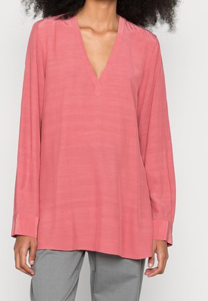 Frau trägt eine lockere, langärmelige rosa Bluse mit tiefem V-Ausschnitt, kombiniert mit grauen Hosen, vor einem schlichten Hintergrund.