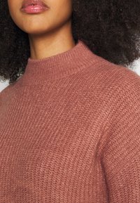 Stickad tröja i dammros med hög krage och texturerad yta. Har en nära passform och mjukt, mysigt tyg.