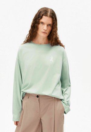 Personne aux longs cheveux bouclés portant une chemise à manches longues vert clair et un pantalon beige, debout avec une main dans la poche.