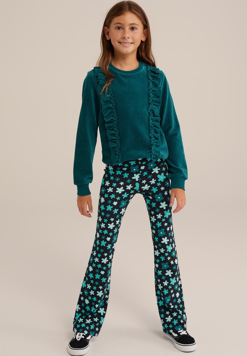 Teal korduroj trenirka s volanima sprijeda, u kombinaciji s crnim širokim hlačama s uzorkom metvica cvjetova i crnim tenisicama.
