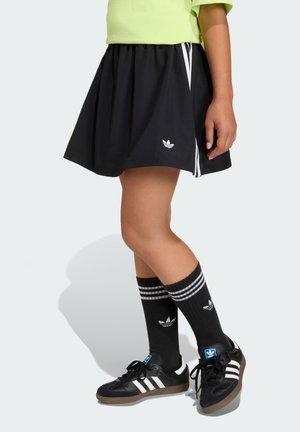 Falda plisada negra con rayas blancas laterales, combinada con calcetas negras hasta la rodilla con rayas blancas y un logo. Zapatillas negras clásicas.