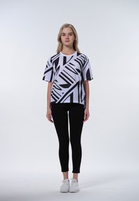 Camicia bianca a maniche corte con un motivo geometrico nero, abbinata a leggings neri e sneakers bianche. Design minimalista, vestibilità informale.
