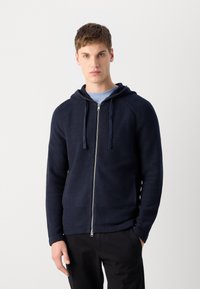 Felpa con zip con cappuccio di colore blu navy, maniche raglan e cordino. Presenta una finitura testurizzata e una zip metallica. Abbinata a pantaloni neri.