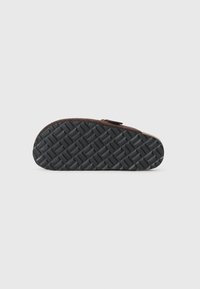 Brown suede slipper with a textured black rubber outsole, featuring a curved shape and a subtle fastening detail.
