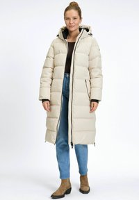 Vrouw in een lange crèmekleurige pufferjas met capuchon, blauwe jeans, bruine laarzen en zwarte top, staande tegen een effen witte achtergrond.