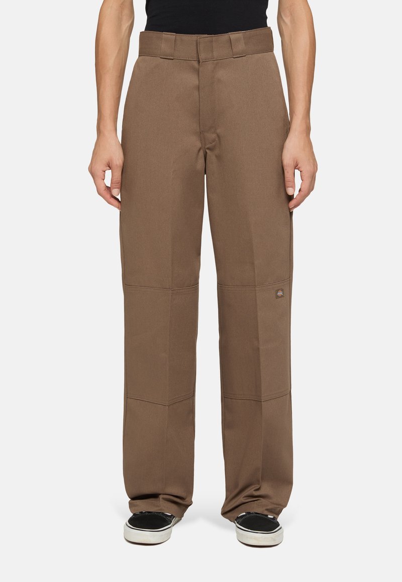 Pantaloni larghi di colore marrone realizzati in un tessuto resistente. Presentano pannelli cuciti e una toppa con logo vicino all'orlo. Indossati con sneakers nere.