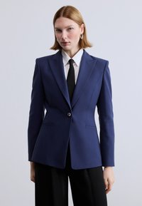 Ναυτικό μπλέ blazer με δομημένο σχέδιο, κούμπωμα με ένα κουμπί, αιχμηλούς πέτο και δύο μπροστινές τσέπες, φορεμένο πάνω από λευκό πουκάμισο και μαύρη γραβάτα.