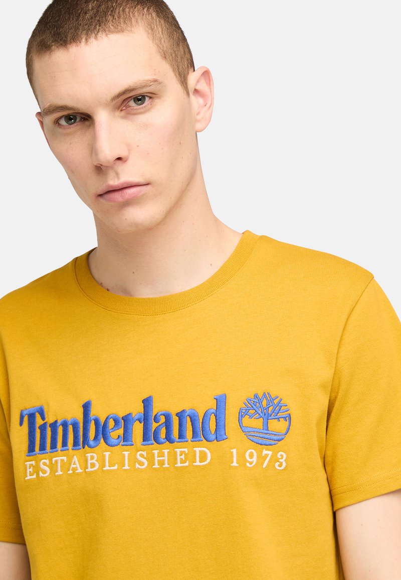 Żółta bawełniana koszulka z niebieskim haftowanym logo "Timberland" oraz grafiką drzewa, z białym napisem "ZAŁOŻONA W 1973 R." poniżej.