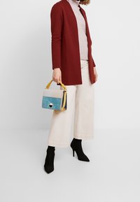 Sac à main multicolore avec une poignée jaune, comprenant des sections bleues, crème et marine. Accessoirisé avec un cardigan côtelé bordeaux et un pantalon large crème.